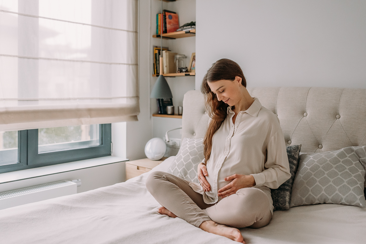 mama w ciąży siedzi na łóżku i pokazuje swój brzuszek