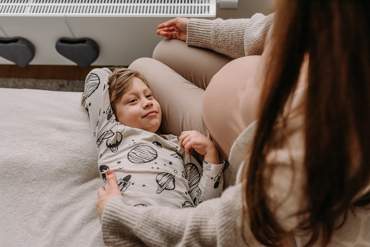 synek leży z mamą na łóżku i patrzy na jej ciążowy brzuszek