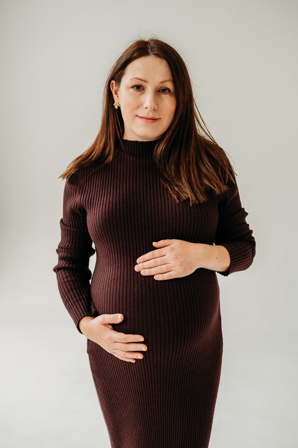 kobieta w ciąży stoi na białym tle i pozuje do zdjęć w bordowej sukience 