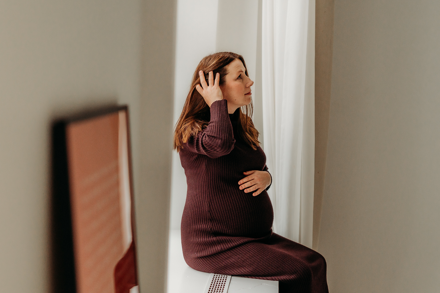 dziewczyna w ciąży w warszawie siedzi na parapecie w bordowej sukience i pozuje do zdjęć