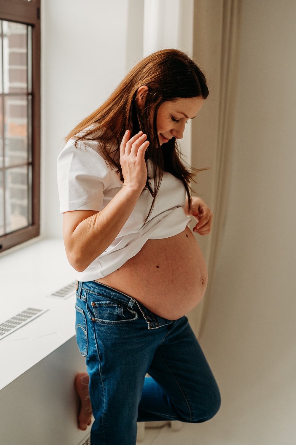 dziewczyna w ciąży siedzi na parapecie i podnosi koszulkę pokazując ciążowy brzuszek