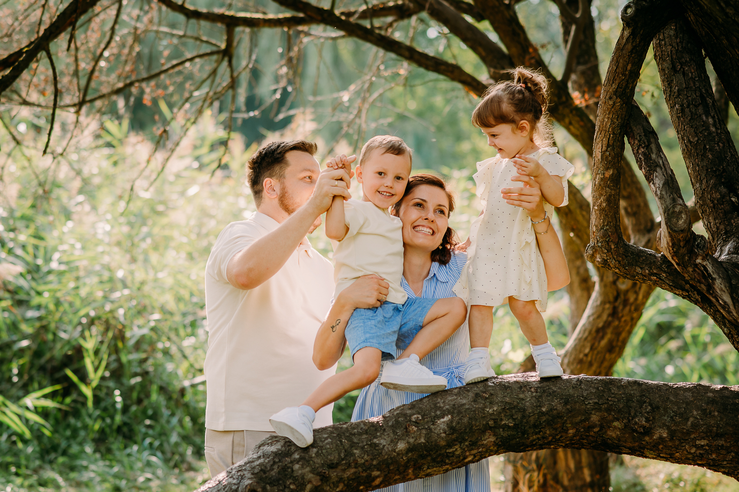 rodzina stoi przy drzewie, rodzice gilgoczą dzieci, wszyscy się śmieją, tak może wyglądać sesja z dziećmi w warszawie