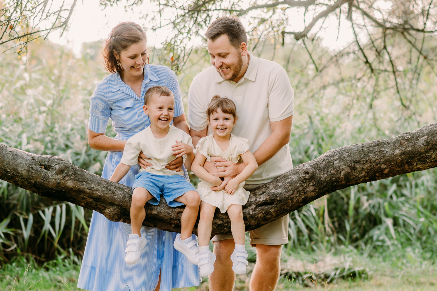 rodzina stoi przy drzewie, rodzice gilgoczą dzieci, wszyscy się śmieją, tak może wyglądać sesja z dziećmi w warszawie